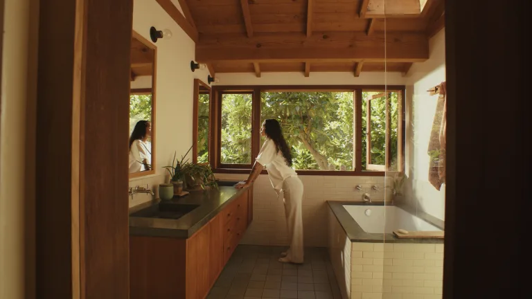 A woman in a sunlit wooden bathroom leaning at the counter, looking out to a garden window.